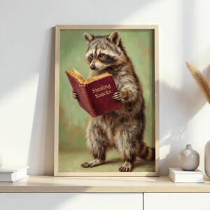 May include: A framed print featuring a raccoon reading a book with the text "Stealing Snacks." The raccoon is depicted with detailed fur and is set against a green background. The book is a deep red color.
