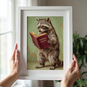 May include: A framed art print featuring a raccoon reading a book with the text "Stealing Snacks." The raccoon is depicted in a realistic style with detailed fur and is set against a green background. The frame is white.