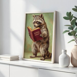 May include: A framed art print featuring a raccoon reading a book with the text "Stealing Snacks." The raccoon is depicted in a realistic style with a green background. The frame is white.