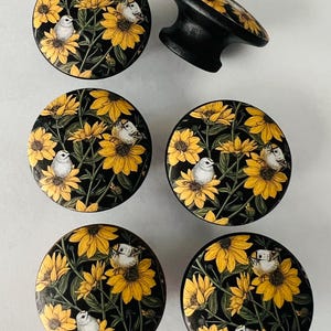 May include: Set of six decorative cabinet knobs. Each knob features a black background with a pattern of yellow sunflowers and small white birds. One knob is a pull handle. The knobs are made of wood.