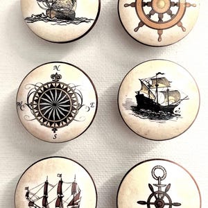 May include: Six round ceramic knobs with nautical designs. The designs include a ship with sails, a compass, a ship's wheel, and an anchor with a ship's wheel.
