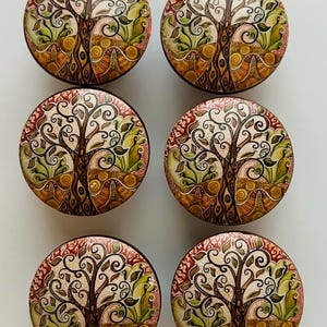 May include: Six round decorative cabinet knobs with a tree of life design. Each knob features a detailed illustration of a tree with swirling branches, leaves, and roots in shades of brown, green, and orange. The knobs have a dark brown border.