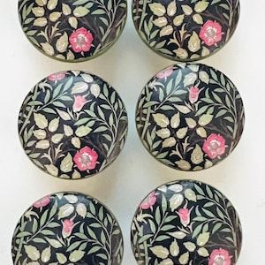 May include: Six round cabinet knobs with a floral design. The knobs feature a black background with pink roses, green leaves, and light green foliage. The knobs are likely made of ceramic or glass.