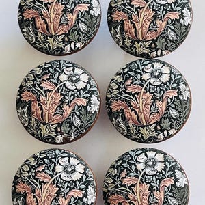 May include: Six round cabinet knobs with a floral design. The knobs feature a black background with a pattern of white flowers, peach-colored leaves, and green foliage. The edges of the knobs are brown.
