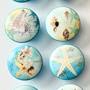 May include: Six round, blue-rimmed, decorative knobs with a variety of beach-themed designs. The designs include seashells, starfish, and handwritten text. The text on one knob reads "You Light Up My Life".