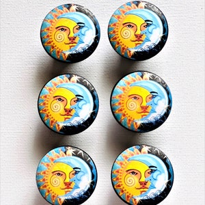 May include: Six round, wooden knobs with a sun and moon design. The sun is yellow with a spiral design and a face. The moon is blue with a face. The background is black with white stars.