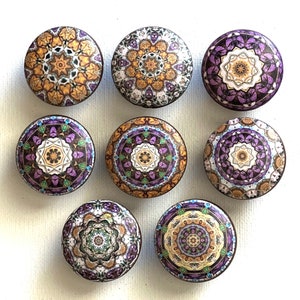 May include: Eight round decorative knobs with intricate mandala designs in shades of purple, gold, and white. The knobs have a glossy finish and are perfect for adding a touch of bohemian style to furniture or cabinets.