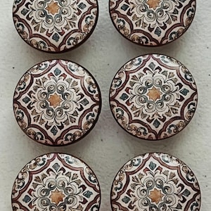 May include: A set of six round cabinet knobs featuring a symmetrical, intricate pattern in shades of brown, cream, blue, and yellow. Each knob displays a central star motif surrounded by ornate, swirling floral-inspired decorative elements.