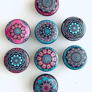 May include: A collection of eight round decorative cabinet knobs. Each knob features a unique, colorful mandala design in shades of pink, blue, and black. The knobs are arranged on a white surface.