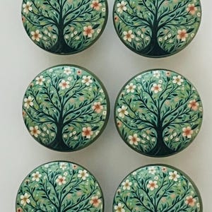 May include: A set of six round cabinet knobs, each featuring a hand-painted design of a dark tree with green leaves and small white and pink flowers. The knobs are circular with a smooth finish, displaying a consistent botanical illustration.