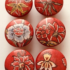 May include: Six round red wooden knobs with floral patterns. The knobs have a variety of floral designs in shades of white, yellow, and red.
