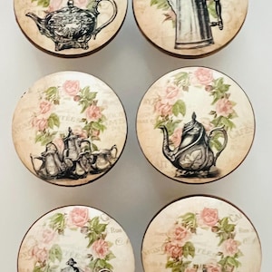 May include: Set of six decorative cabinet knobs with vintage-style illustrations. Each knob features a different black and white teapot or coffee pot design, surrounded by a wreath of pink roses on a cream background. The knobs have a dark brown edge.