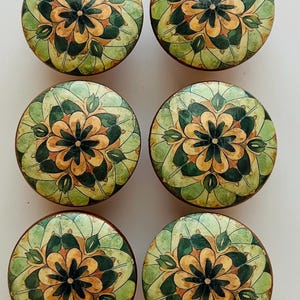May include: Six round cabinet knobs with a floral design. The knobs feature a central flower in shades of brown and yellow, surrounded by green and cream-colored leaf patterns. The knobs are likely made of wood.