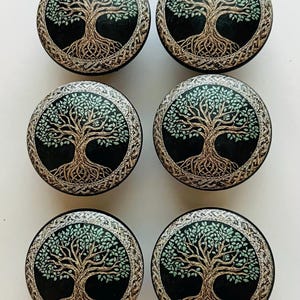 May include: Six round cabinet knobs with a Tree of Life design. Each knob features a detailed tree illustration with a brown trunk and green leaves set against a black background. The knobs are framed by a silver Celtic knot border.