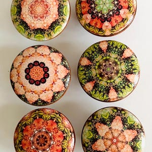 May include: Six round decorative cabinet knobs with floral mandala designs. The knobs feature a variety of colors, including pink, orange, green, and brown. Each knob has a glossy finish and a wooden base.