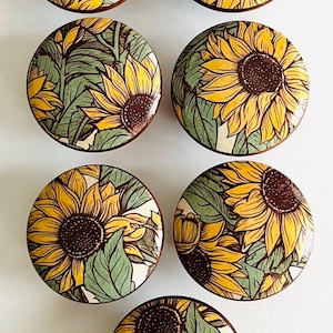 May include: Six round decorative cabinet knobs with a sunflower design. Each knob features a yellow sunflower with a brown center and green leaves against a white background. The knobs have a brown wooden edge.