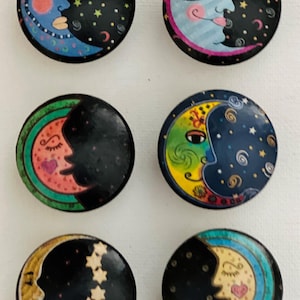 May include: Six round, colorful, decorative buttons with a crescent moon design. Each button features a different color scheme and design, including blue, green, yellow, pink, and black.