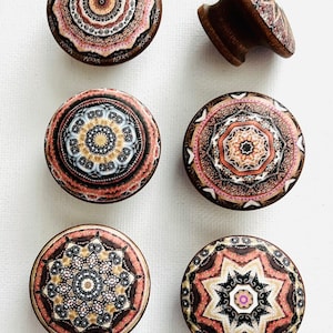 May include: Six round wooden knobs with intricate mandala designs in shades of brown, pink, and white. The knobs have a raised edge and a smooth, polished finish.