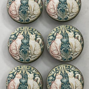 May include: Six round decorative cabinet knobs. Each knob features a design of two cats sitting side-by-side, set against a backdrop of teal and cream-colored floral patterns. The knobs have a metallic, bronze-colored edge.