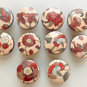 May include: A collection of ten round cabinet knobs with floral and bird designs. The knobs feature a cream-colored background with red, white, and green botanical and avian illustrations. Each knob has a dark brown edge.