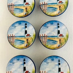 May include: Six round wooden knobs with a blue border and a painted image of a lighthouse on a beach. The lighthouse is black and white with a red roof. The beach is yellow with blue water and a wooden fence.