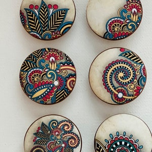 May include: Six round wooden coasters with a cream background and colorful paisley designs. The designs feature swirls, leaves, and flowers in shades of red, blue, yellow, and black.