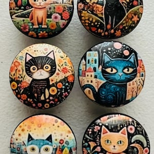 May include: Six round decorative cat-themed buttons. Each button features a different cat illustration with colorful floral and cityscape backgrounds. The cats are depicted in various colors, including orange, black, and blue.