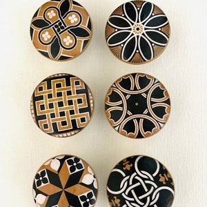 May include: Six round, wooden knobs with black, white, and gold geometric patterns. The knobs are approximately 1.5 inches in diameter.