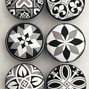 May include: A set of six circular cabinet knobs featuring distinct black, white, and gray geometric patterns. Each knob displays a unique design, including floral motifs, star-like shapes, and intricate lattice work on a smooth, round surface.