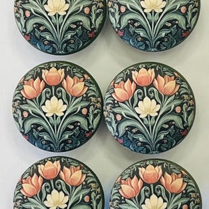 May include: A set of six circular cabinet knobs, each featuring a dark background with a repeating botanical illustration of light orange and white tulips, green leaves, and small red accents arranged in a symmetrical floral pattern.