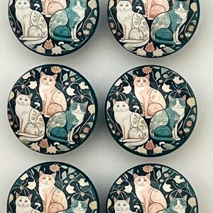 May include: A set of six circular cabinet knobs featuring a dark teal background with a repeating pattern of three cats in white, peach, and blue-green tones, interspersed with small white and peach floral elements.