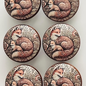 May include: A set of six round wooden cabinet knobs, each featuring a detailed illustration of an adult fox and a small kit nestled among foliage and flowers in shades of orange, brown, and cream with a dark wood border.