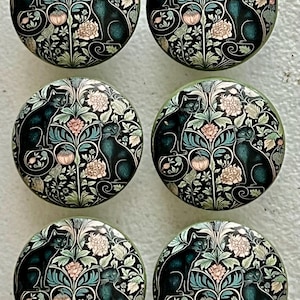 May include: Six round cabinet knobs with a black cat design. The knobs feature a black background with floral and cat motifs in shades of pink, teal, and cream. The knobs are likely made of ceramic or glass.