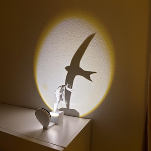 Puede incluir: Una lámpara de noche blanca proyecta una silueta de pájaro sobre una pared. La lámpara está sobre una mesita de noche blanca junto a una cama. La sombra del pájaro está en un círculo de luz amarilla. Una segunda lámpara está en la mesa.