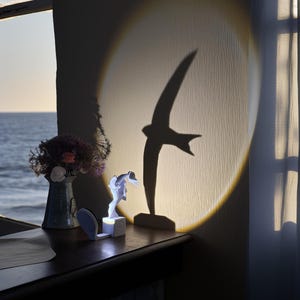 Könnte beinhalten: Eine kleine, weiße Vogelskulptur wird beleuchtet und wirft einen großen Vogelschatten an die Wand. Der Schatten befindet sich in einem Scheinwerfer. Eine Vase mit Blumen steht auf einer Holzoberfläche in der Nähe eines Fensters mit Meerblick.