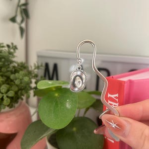 May include: A silver metal bookmark featuring a cowboy hat charm. The hat charm has a detailed brim and central design. The bookmark is held near a pink book and green plants.