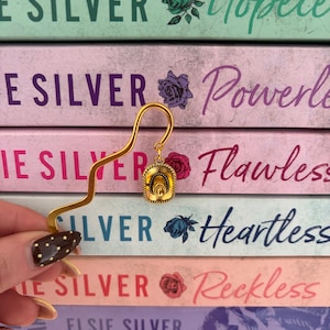 May include: A gold-coloured metal bookmark with a cowboy hat charm, placed on a stack of books. The books are titled "Hopeless", "Powerless", and "Flawless" by Elsie Silver. The background features pastel colours.