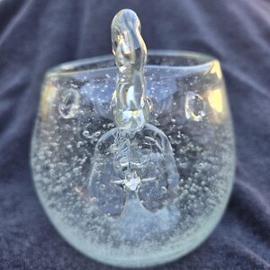 Peut inclure: Un verre transparent avec une poignée décorative. La coupe est remplie de petites bulles, ce qui lui donne un aspect texturé. La poignée est sculptée dans une forme unique, ajoutant une touche artistique à la verrerie.