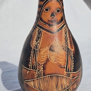 Puede incluir: Una pieza de arte de calabaza hecha a mano con un diseño tallado. La calabaza es marrón con acentos marrón oscuro y presenta una cara tallada y una figura que sostiene una bola. El diseño incluye patrones geométricos y líneas.