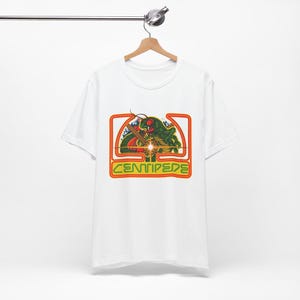 May include: White t-shirt featuring a retro graphic design. The design includes a green centipede with red accents, a yellow laser beam, and the word "CENTIPEDE" in yellow and green. The graphic is framed in a red and orange rectangle.