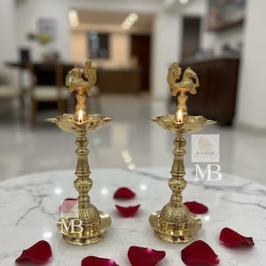 Pode incluir: Duas lâmpadas de óleo de latão dourado ornamentadas com um desenho de pavão, cada uma com um pavio aceso. As lâmpadas estão sobre uma superfície de mármore branco, rodeadas por pétalas de rosa vermelhas. As lâmpadas têm uma base e um caule decorativos.