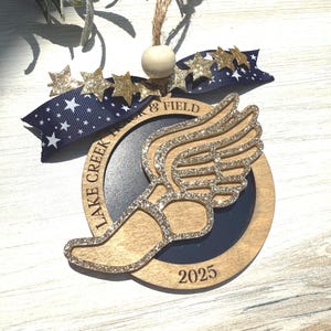 May include: Wooden track and field ornament with a glittery winged foot design. The ornament has a navy blue and gold ribbon with star accents and the text "Lake Creek Track & Field 2025". A wooden bead and twine are attached for hanging.