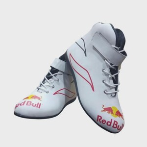 Puede incluir: Un par de zapatillas de carreras blancas de ca&ntilde;a alta con el logotipo de Red Bull en rojo y amarillo en la puntera. Las zapatillas tienen cordones blancos, una tira de velcro en el tobillo y detalles gr&aacute;ficos rojos en los laterales.