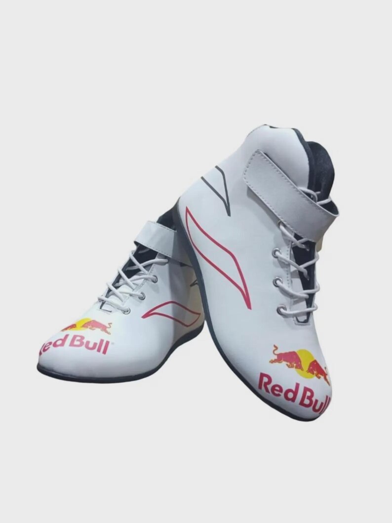 Puede incluir: Un par de zapatillas de carreras blancas de ca&ntilde;a alta con el logotipo de Red Bull en rojo y amarillo en la puntera. Las zapatillas tienen cordones blancos, una tira de velcro en el tobillo y detalles gr&aacute;ficos rojos en los laterales.