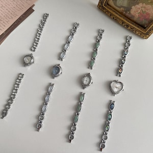 May include: A collection of vintage watches and watch straps. The watches have silver cases and different face designs, including a heart shape. The straps feature decorative oval stones in green, blue, and iridescent colours. The watches are displayed on a white surface.