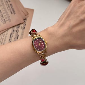 May include: Gold-toned wristwatch with a burgundy face and matching decorative accents. The watch has an oval face with gold hands and markers. The band features decorative red accents. The watch is worn on a wrist.