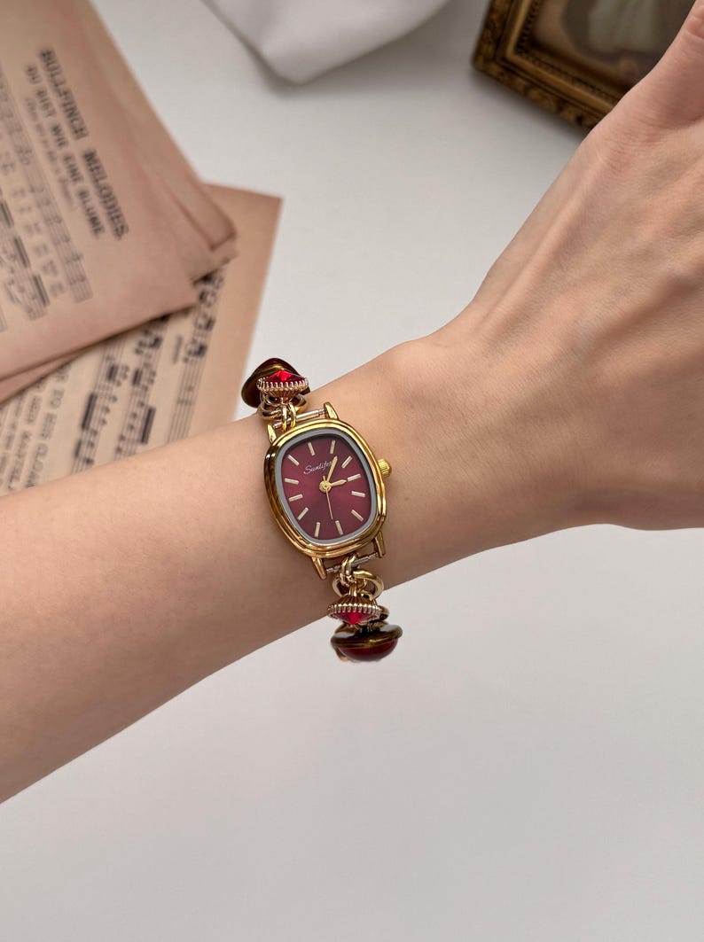 May include: Gold-toned wristwatch with a burgundy face and matching decorative accents. The watch has an oval face with gold hands and markers. The band features decorative red accents. The watch is worn on a wrist.
