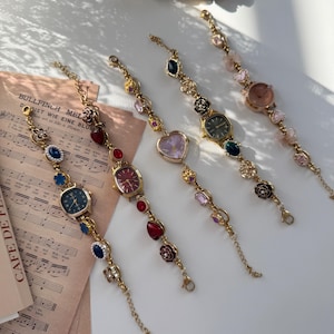 May include: Five gold-toned watch bracelets with various colored gemstones and watch faces. The watch faces are in different shapes, including square and oval. The bracelets are arranged on a white surface with sheet music in the background.