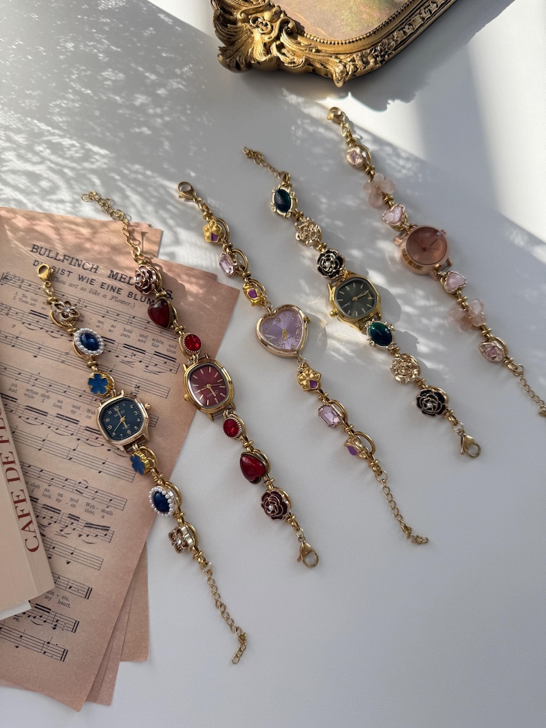 May include: Five gold-toned watch bracelets with various colored gemstones and watch faces. The watch faces are in different shapes, including square and oval. The bracelets are arranged on a white surface with sheet music in the background.
