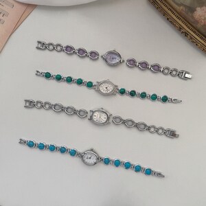 May include: Four silver-toned wristwatches with different colored stone accents. The watches have oval or round faces and matching stone bracelets in purple, green, and turquoise. The watches are displayed on a white surface.